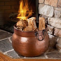 Copper log bucket, filled with chopped firewood, next to a roaring fireplace.