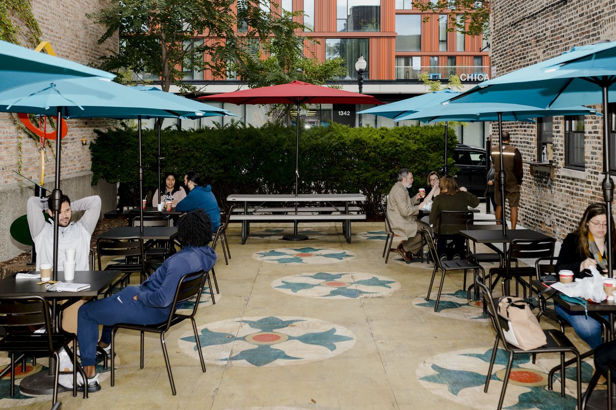  An outdoor patio with a geometric tile pattern floor. Black metal dining tables and chairs are spaced out, shielded from the sun with multi-colored umbrellas.