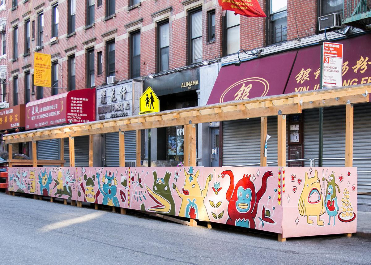  A wooden half wall with a pergola top set on the street in front of a restaurant in New York’s Chinatown. The wooden enclosure is covered with a colorful mural of cute characters.