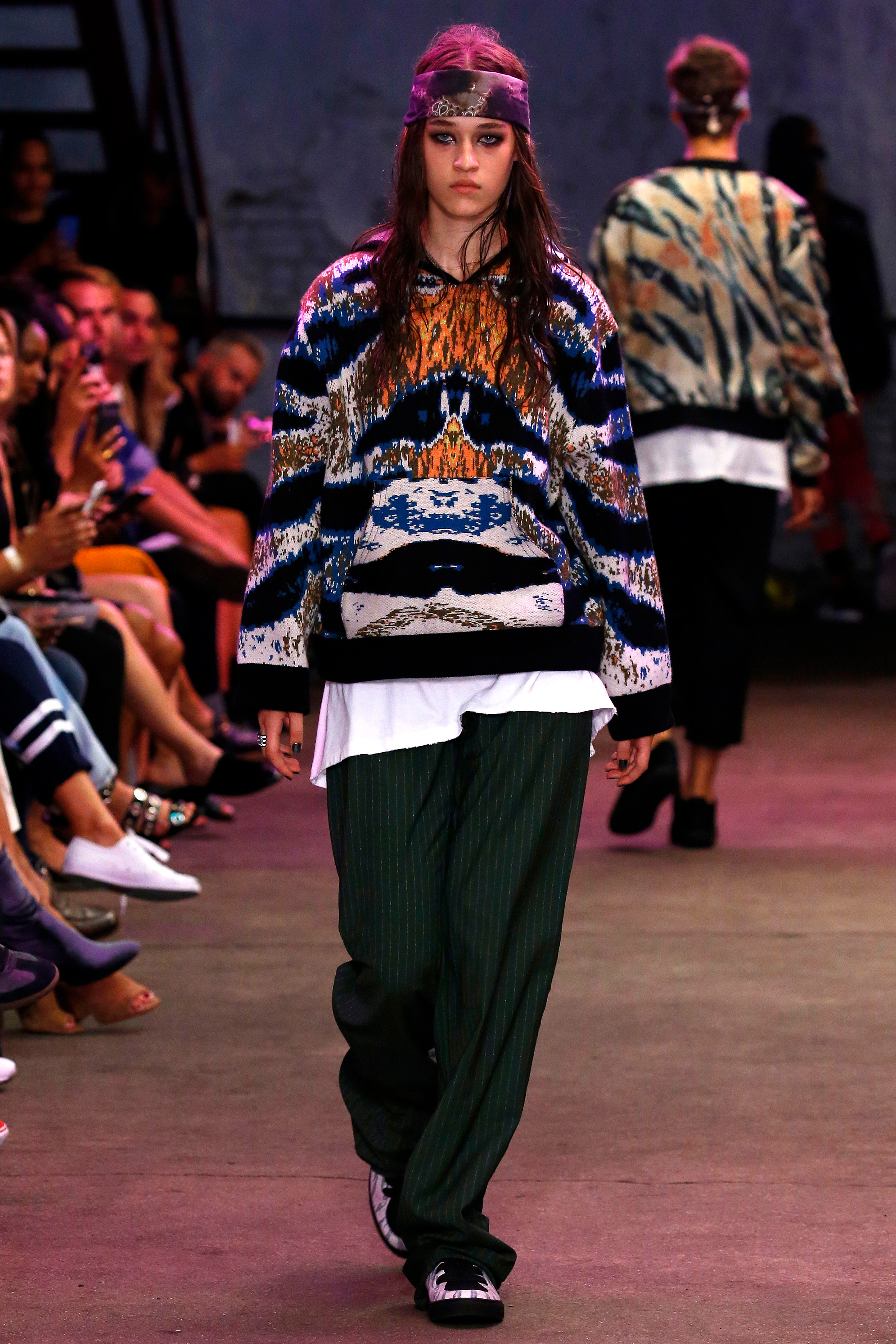 A model in big black pants, a colorful sweater, and a headscarf walks the Baja East runway